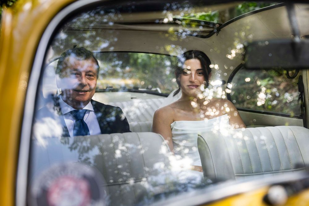 Bride and older man sit in vintage yellow car interior, dappled sunlight through trees reflecting on window.