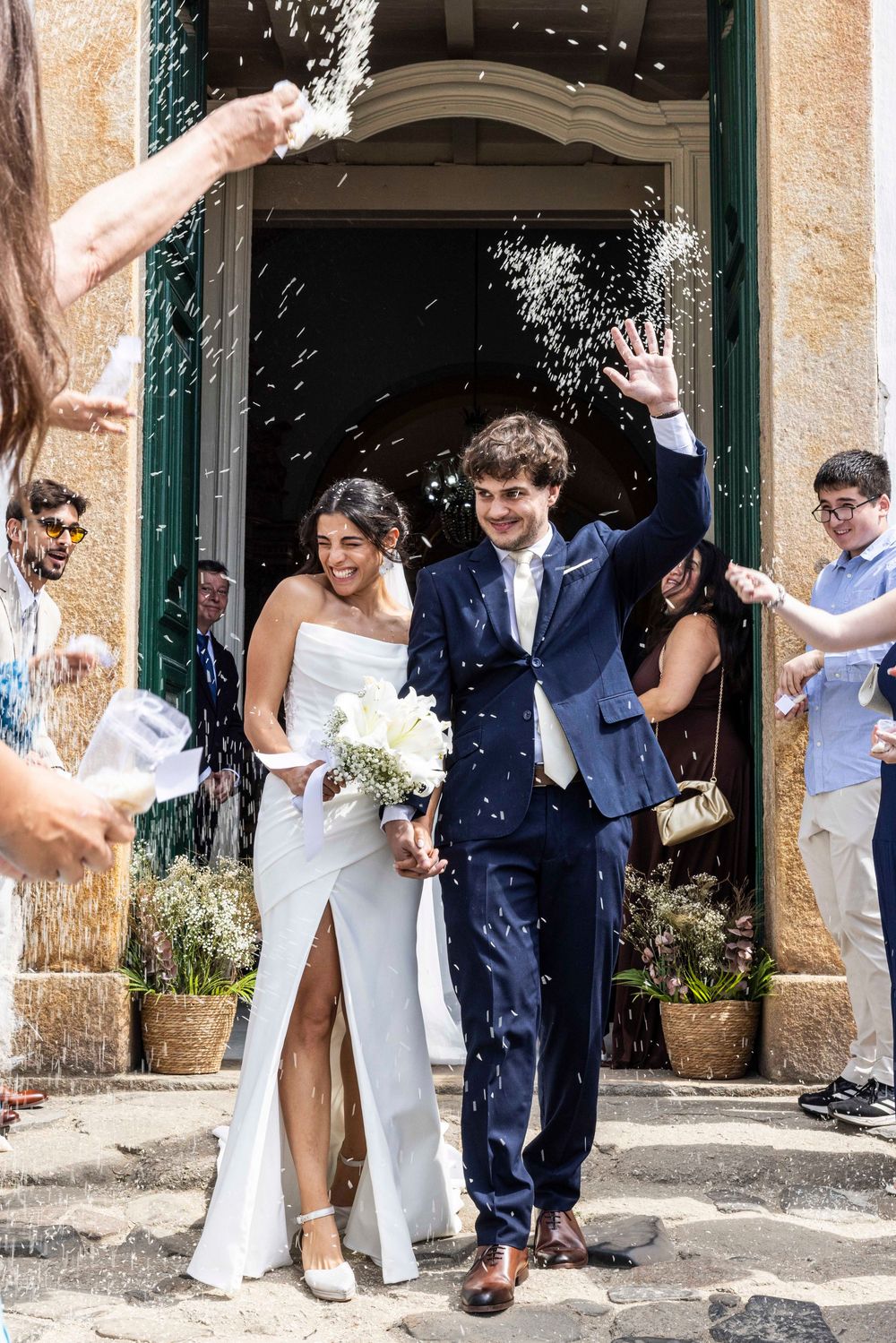 Bride and groom exit church as guests throw rice confetti during joyful outdoor wedding ceremony.