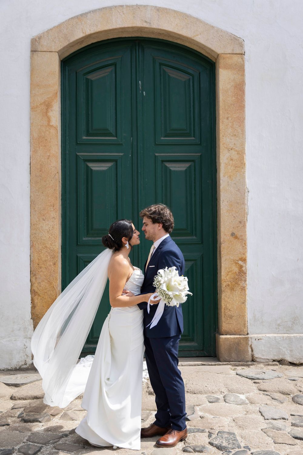 Bride and groom stand facing each other before a green arched door, bride holding white bouquet, veil flowing.