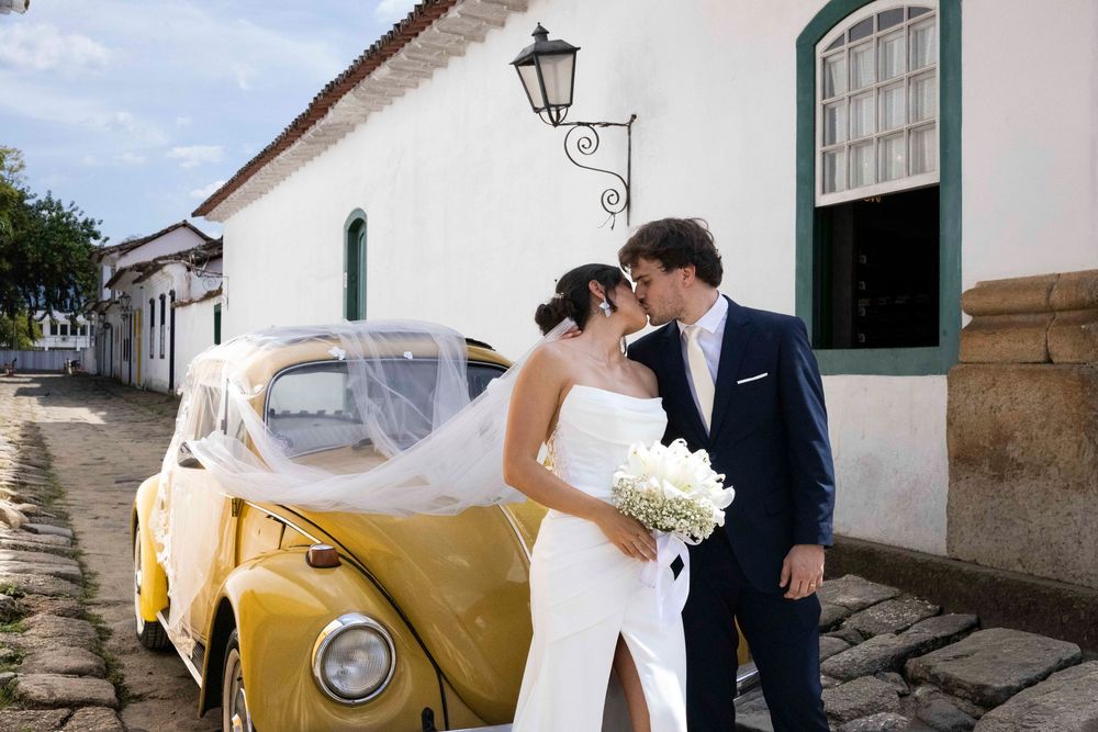 Bride and groom kissing beside a vintage yellow VW Beetle on cobblestone street in historic Brazilian town.