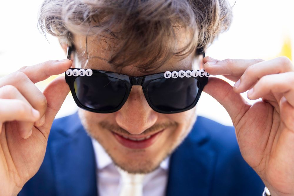 Smiling man in blue suit holding black sunglasses with white letter beads close to his face.