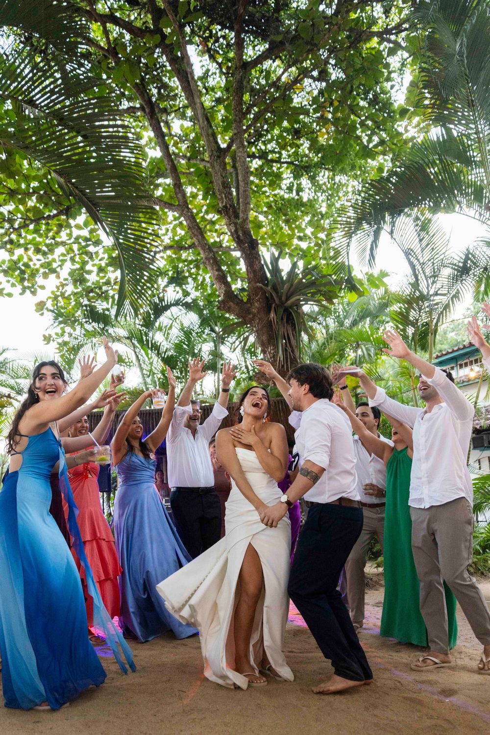 Bride and groom dance joyfully with guests at an outdoor tropical wedding celebration under lush green trees.