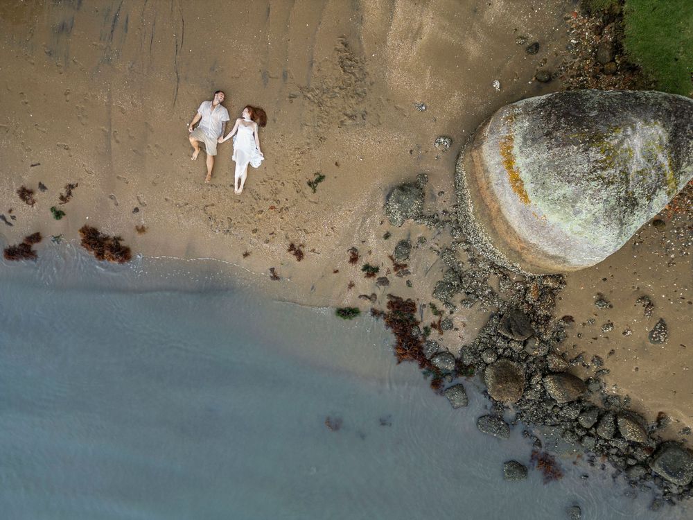 Aerial drone view of couple in white walking hand in hand along sandy beach shoreline.