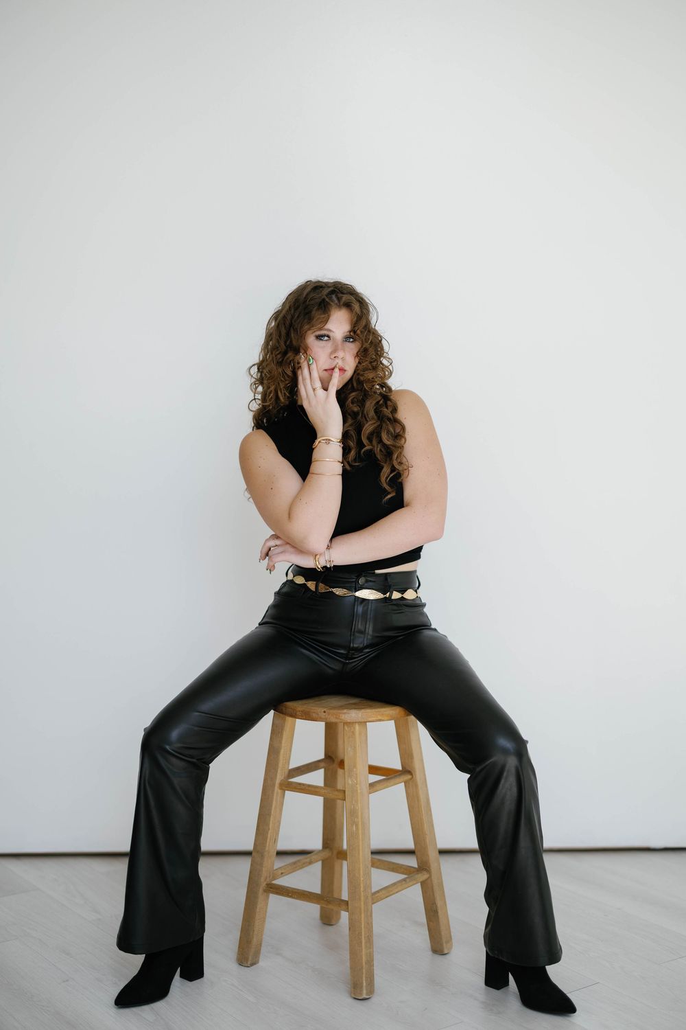 Curly-haired woman in black sleeveless top and leather flare pants posing on wooden stool.