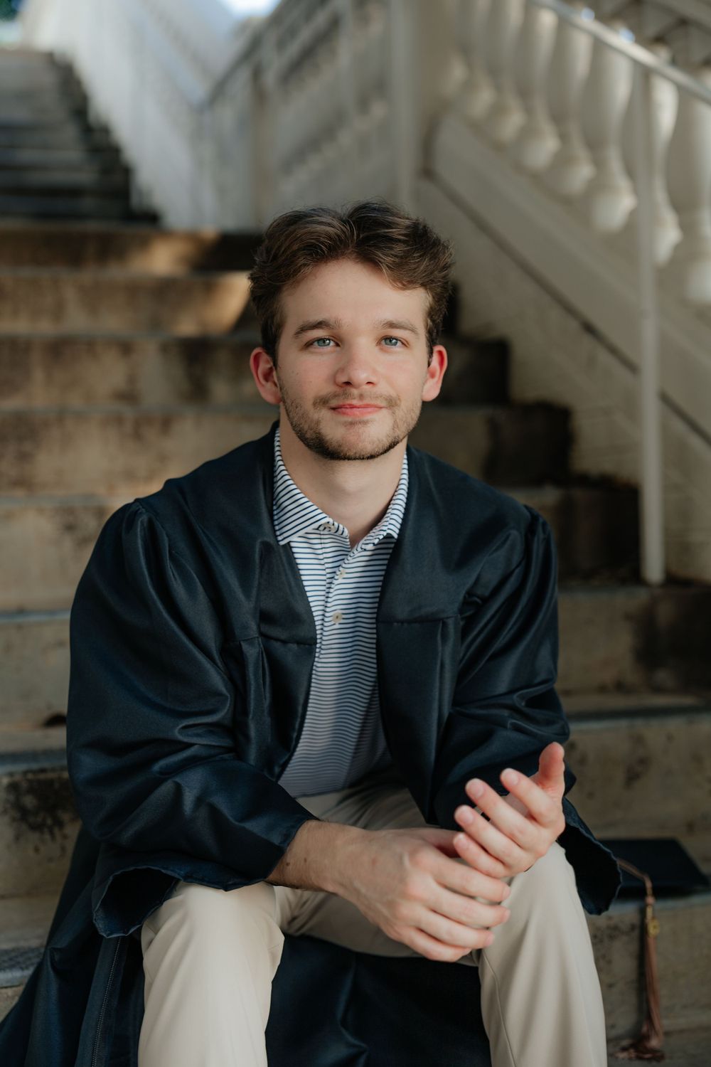 Young man in graduation gown sitting on white stone steps with hands clasped, smiling.