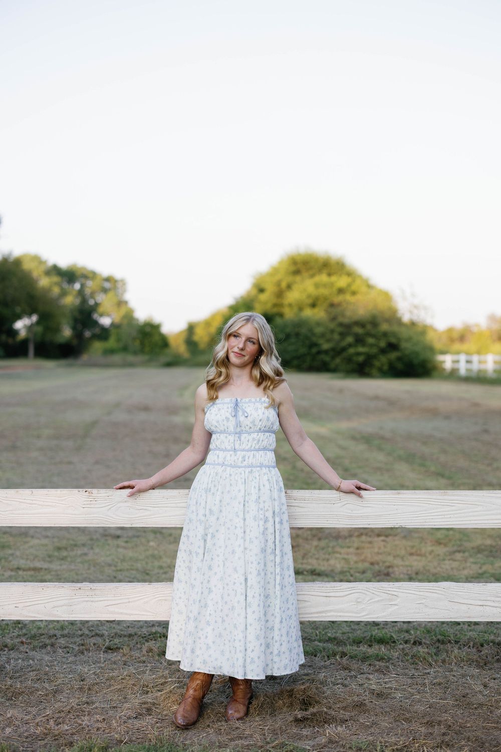 Blonde teen in white floral maxi dress and cowboy boots posing by a wooden fence in a field.