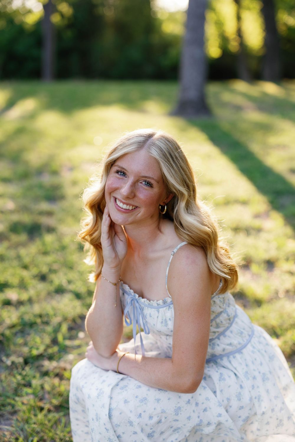 Smiling blonde teenage girl in floral sundress sitting on grass in golden hour sunlight for senior photos.