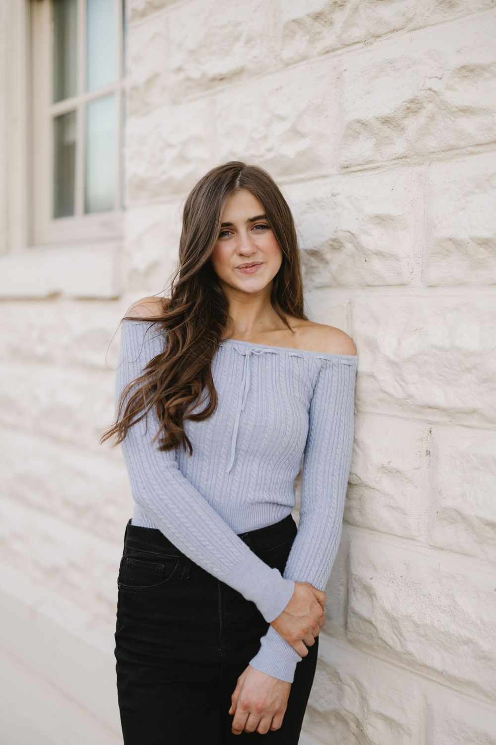 Young woman in light blue off-shoulder ribbed sweater leaning against stone wall.