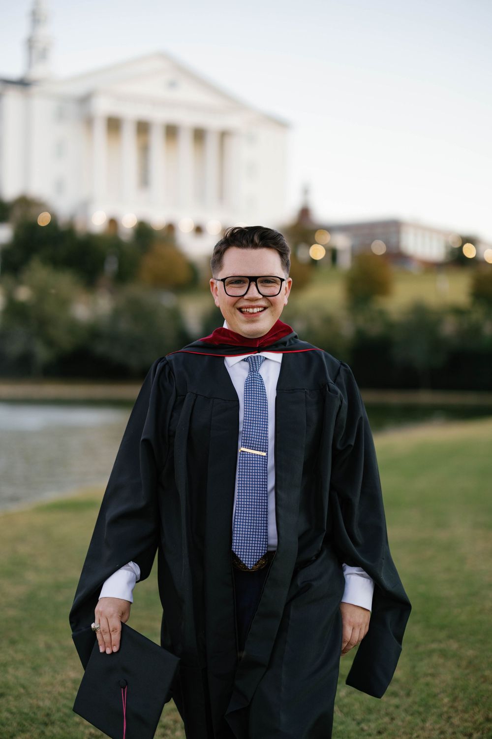 Smiling graduate in black gown and glasses holds cap while standing on green campus lawn.