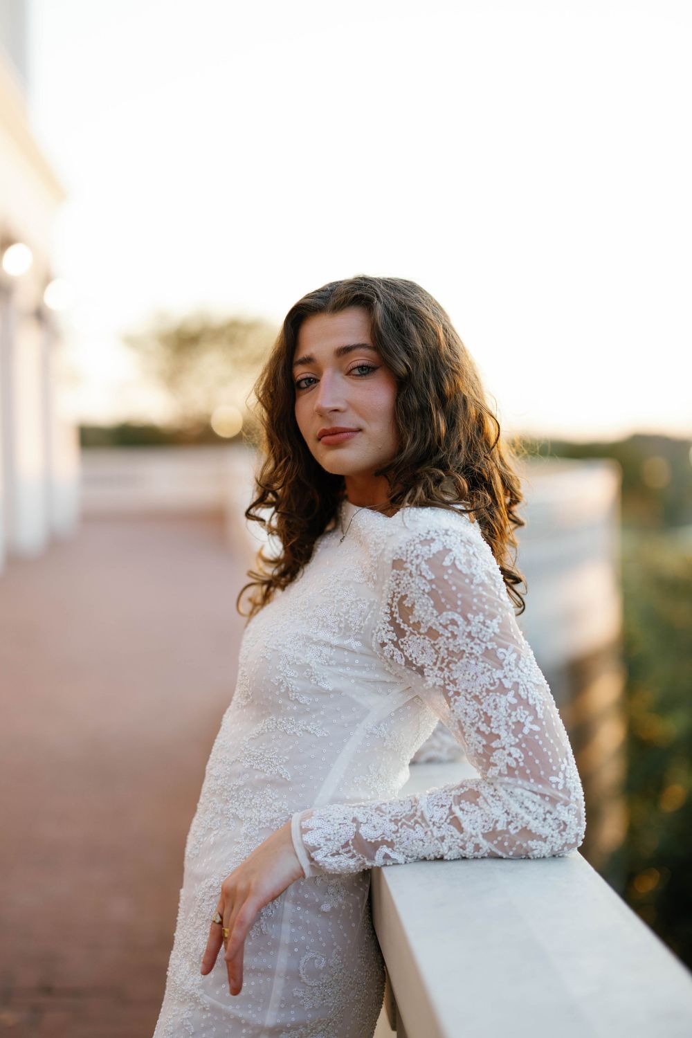 Young woman in white lace long-sleeve dress leans gracefully on railing during golden sunset outdoors.