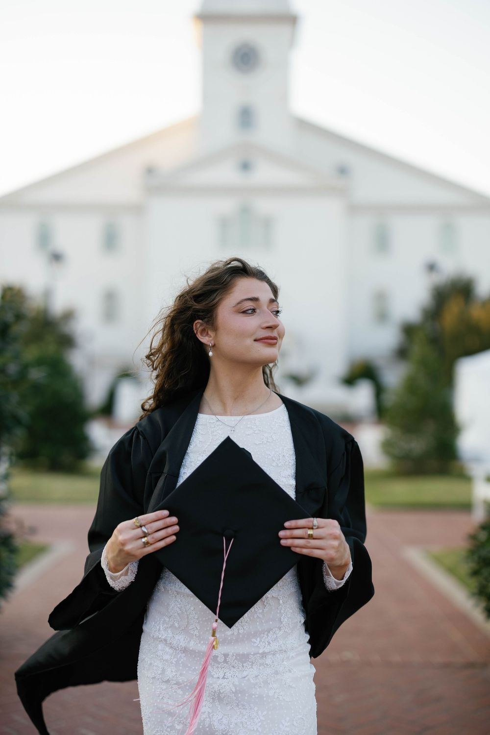 Young woman in graduation gown holds mortarboard cap outside white university chapel building at dusk.