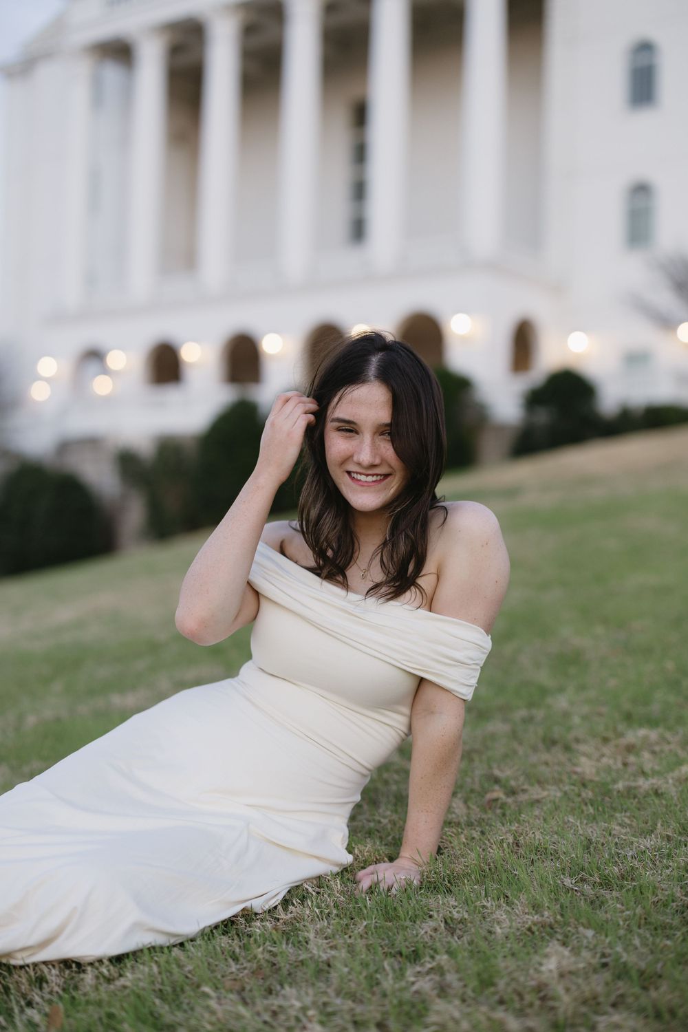 Young woman in elegant off-shoulder cream gown sitting on grass laughing outdoors.