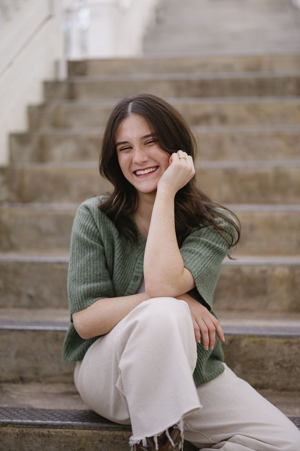 Smiling young woman in green cardigan sits on stone steps with hand resting on chin in casual portrait.
