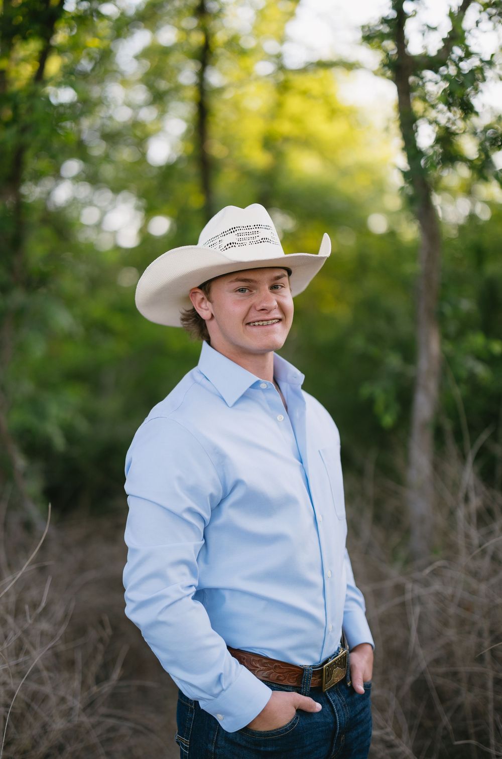 Smiling young man in light blue shirt and white cowboy hat standing in wooded trail.
