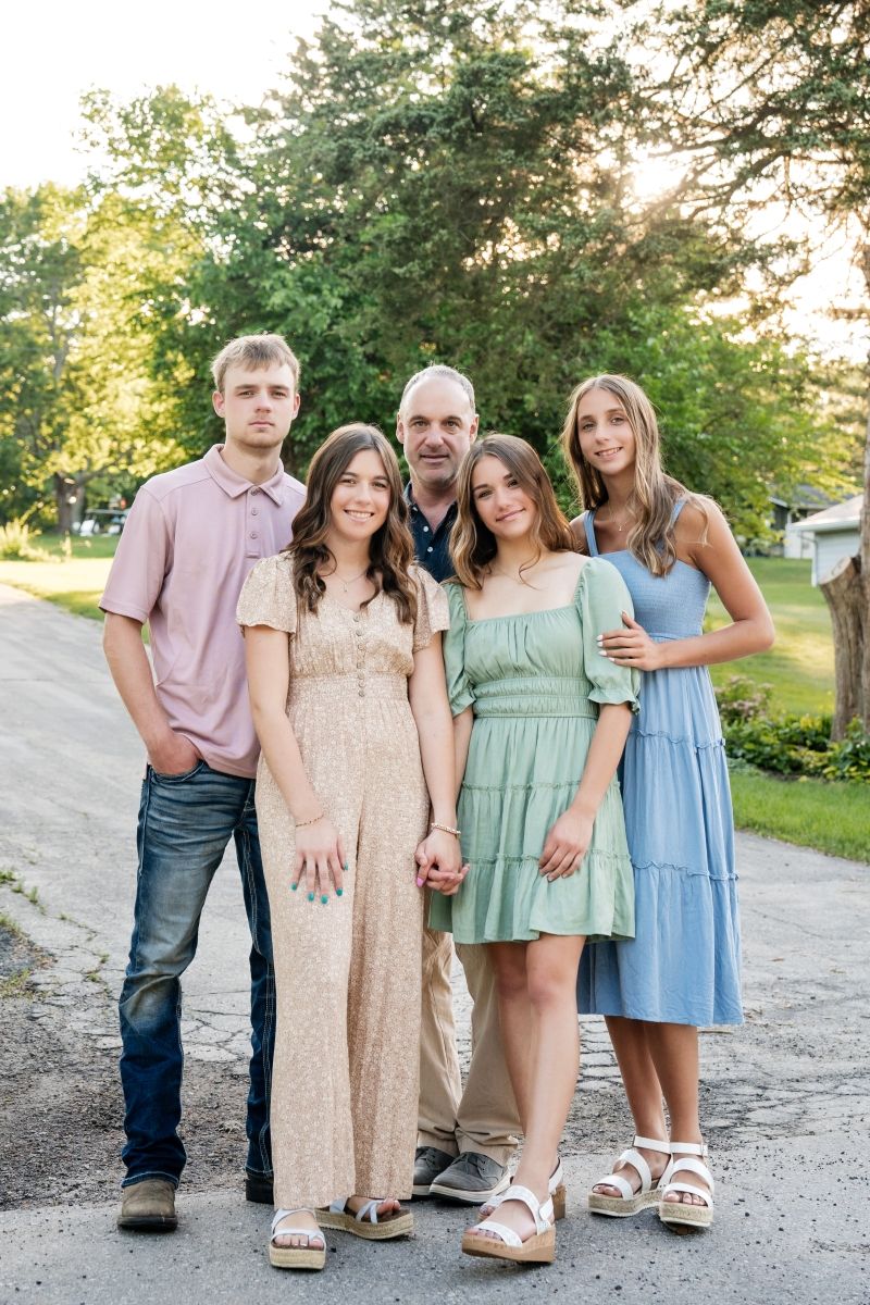 Family photo of family of 5 smiling on a driveway with sunlit trees in background,  shot by Milwaukee family photographer Rachel Dickman