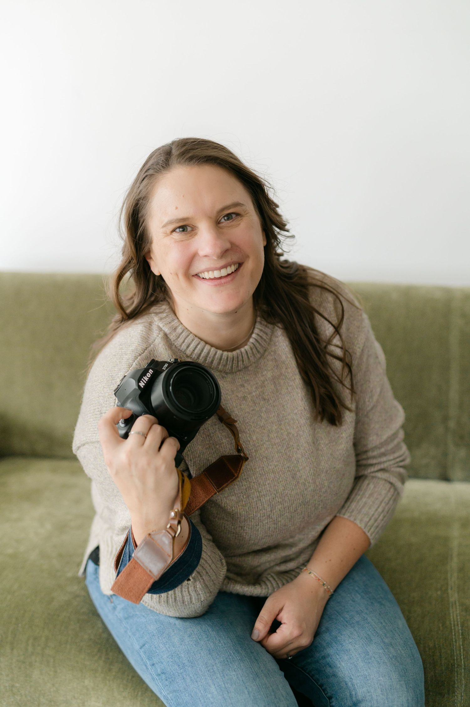 A photographer sits casually on a green couch holding a DSLR camera while wearing a beige sweater and jeans.