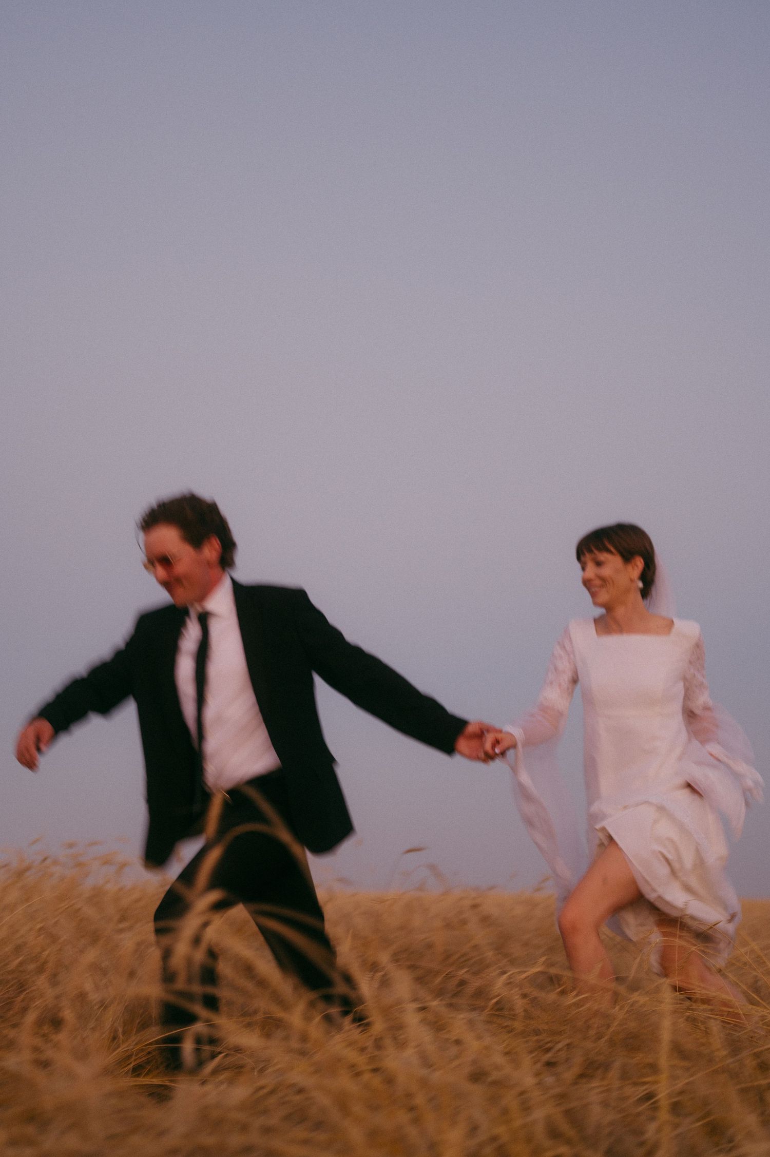 Joyful wedding couple raising joined hands on a rooftop with sunset backdrop.