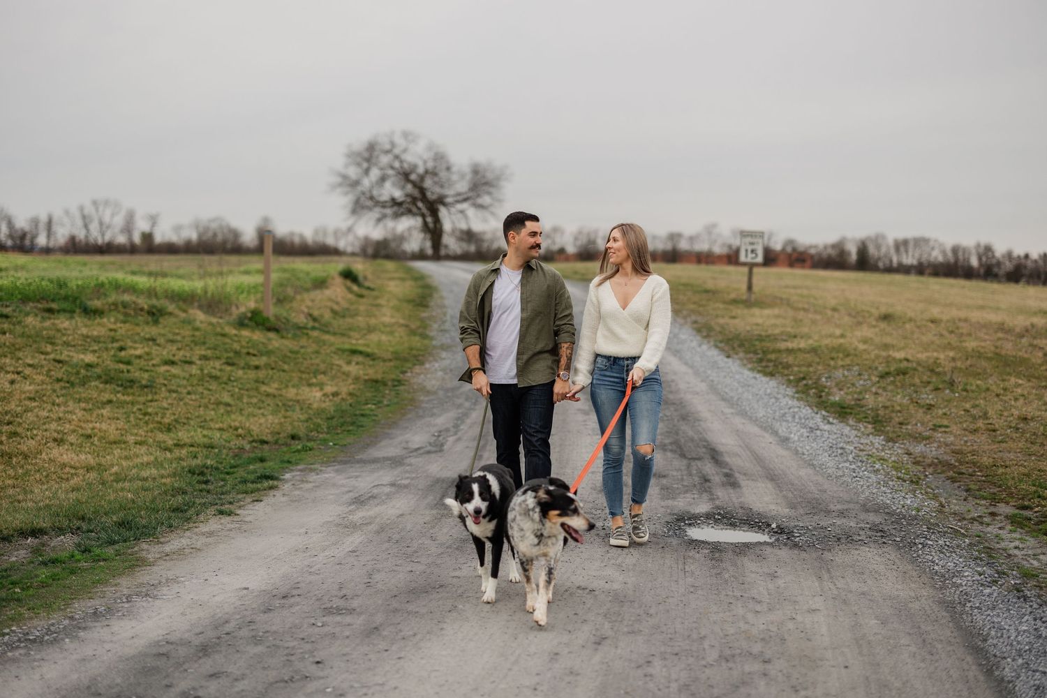 A couple walks their black and white dog down a rural dirt path on a cloudy day.