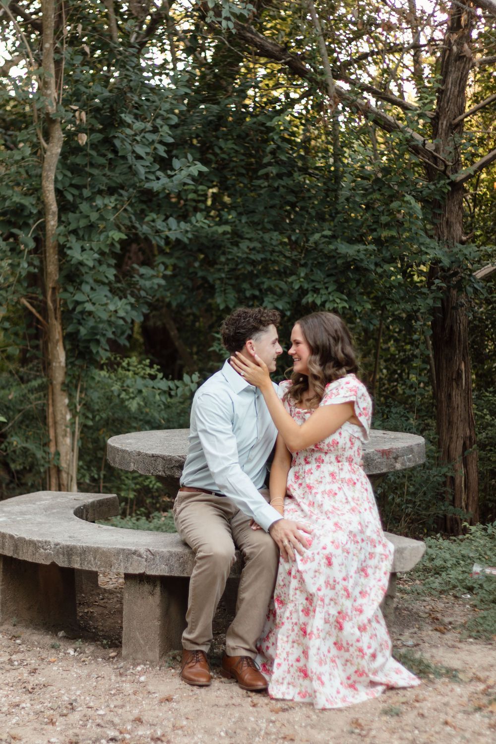 A couple shares a romantic moment on a stone bench surrounded by lush green trees in an outdoor park setting.