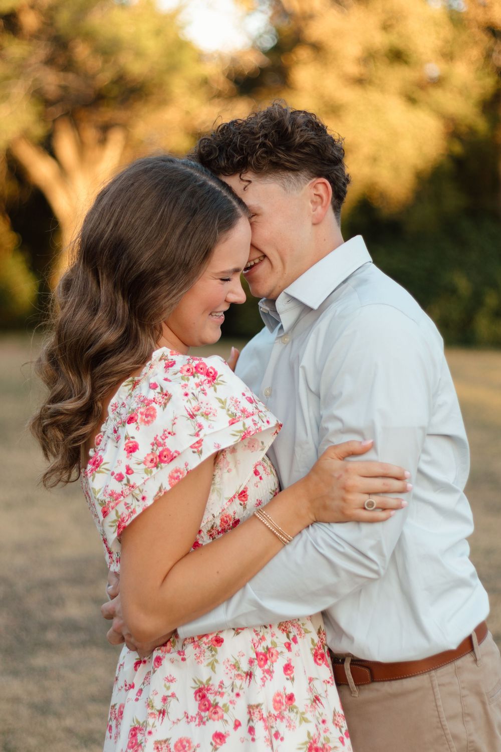 A smiling couple embraces outdoors, woman in floral dress, man in light blue shirt, golden hour lighting.