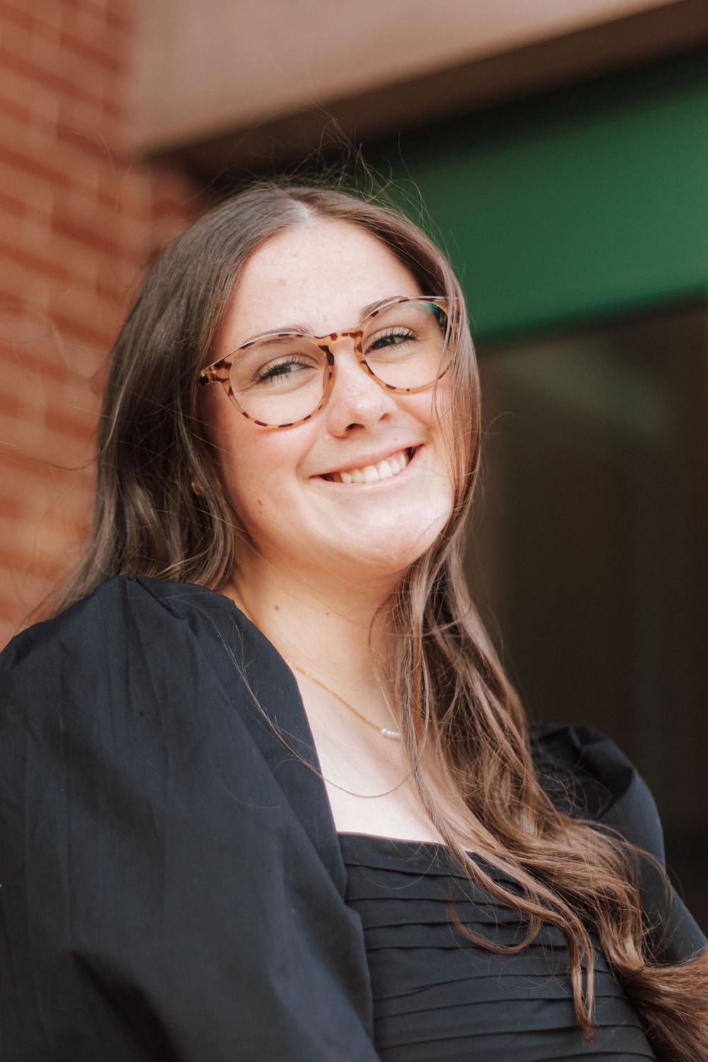 Smiling young woman with long brown hair and tortoiseshell glasses wearing a black graduation gown outdoors.