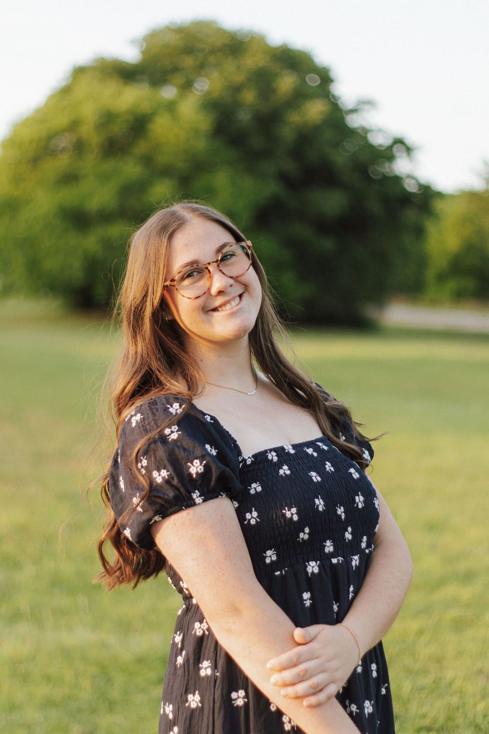 Young woman in floral black dress and glasses smiling outdoors in a sunny green park.