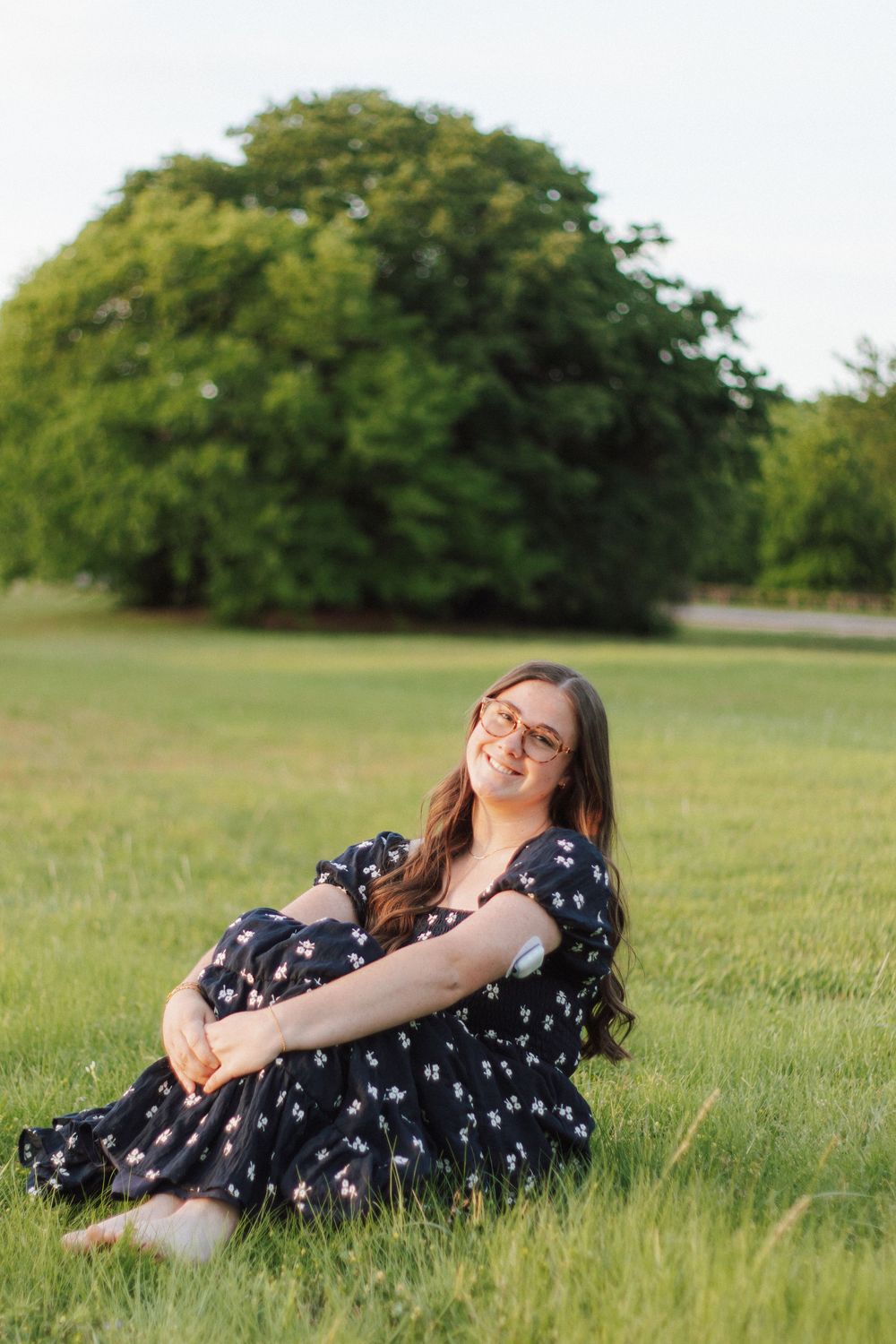 Young woman in floral dress sitting on green grass, smiling outdoors with large tree in background.