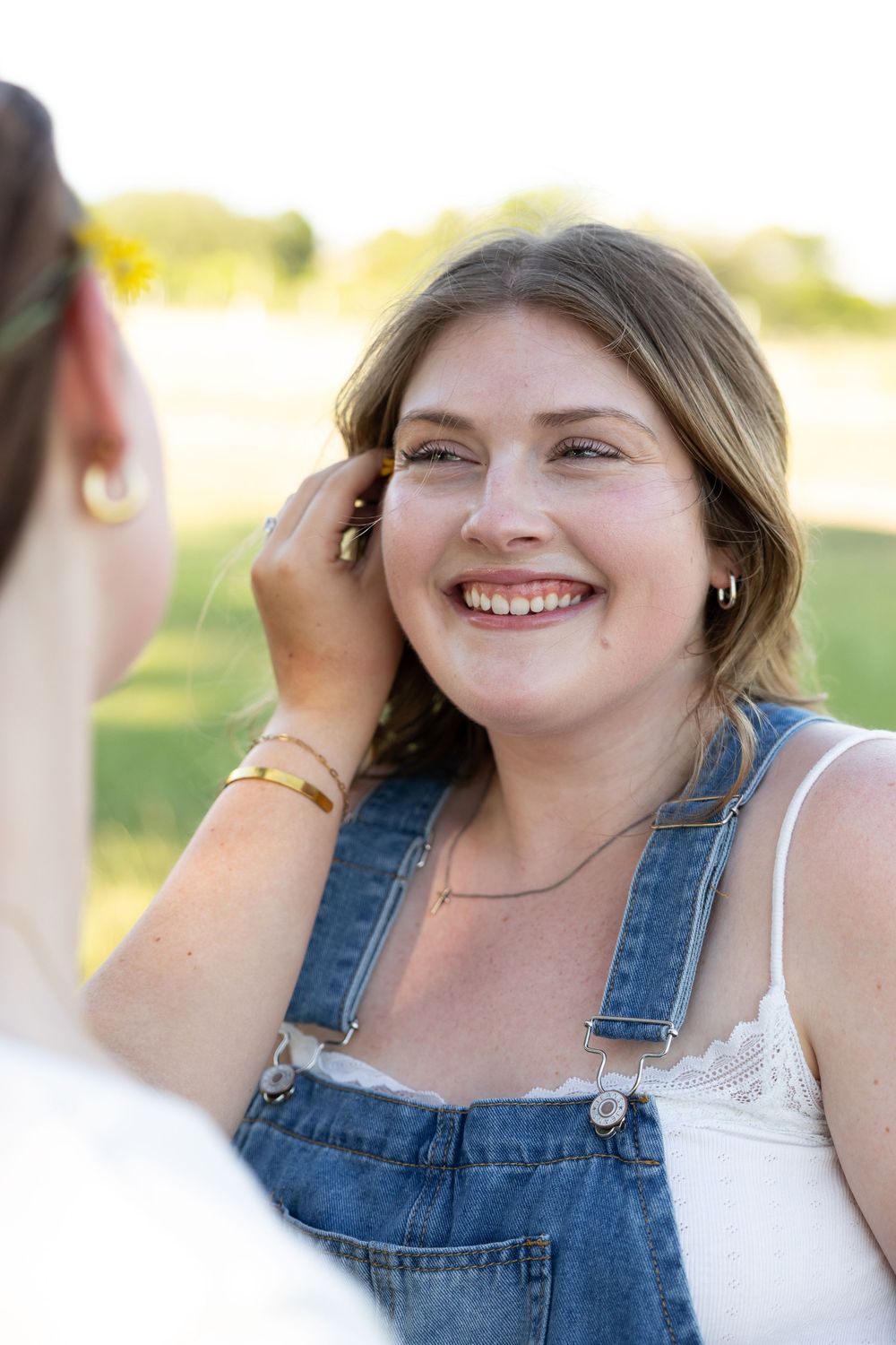 Smiling young woman in denim overalls outdoors, having a flower tucked behind her ear by a friend.