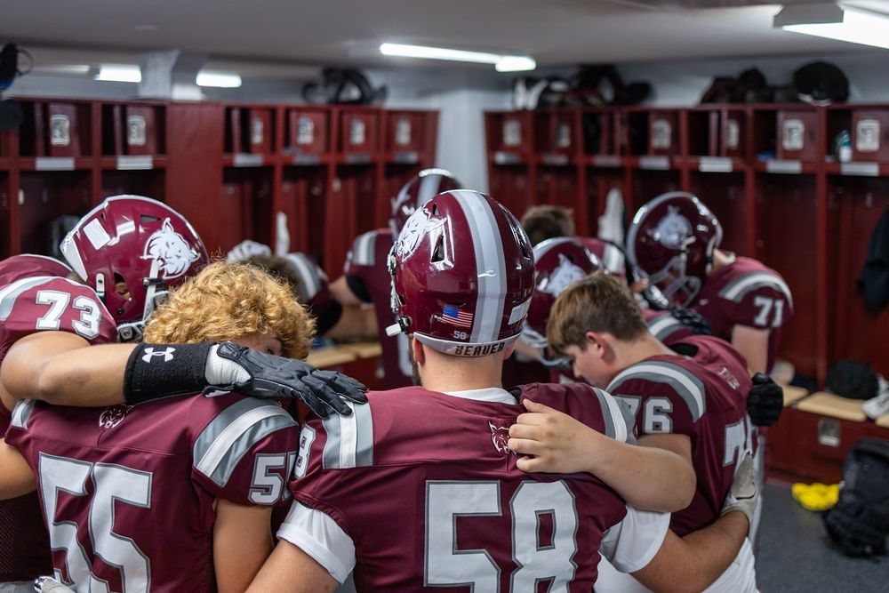 Football team members gather in the locker room wearing maroon uniforms before a game.