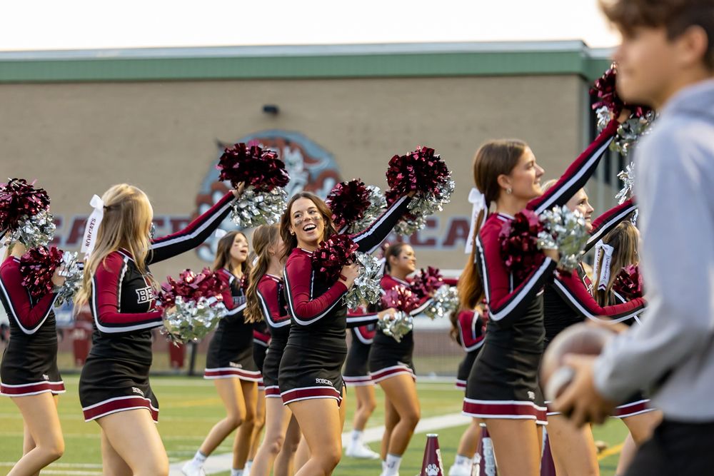 Cheerleaders perform synchronized dance routine in black and red uniforms on a football field sideline.