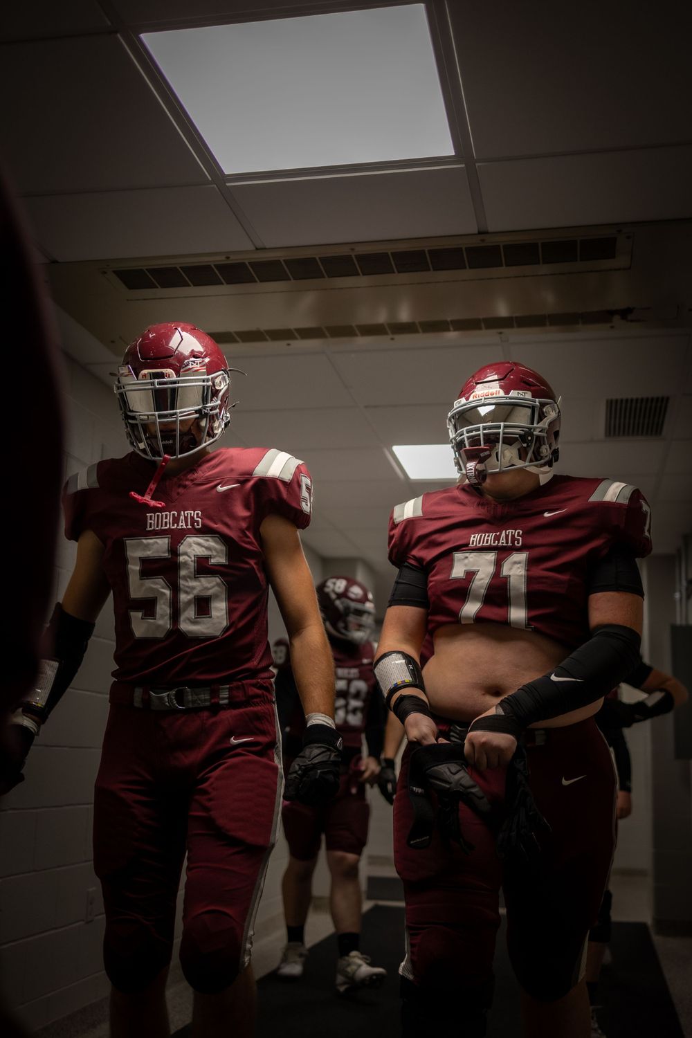 Two football players in red uniforms with numbers 56 and 71 standing in a dark hallway under fluorescent lighting.