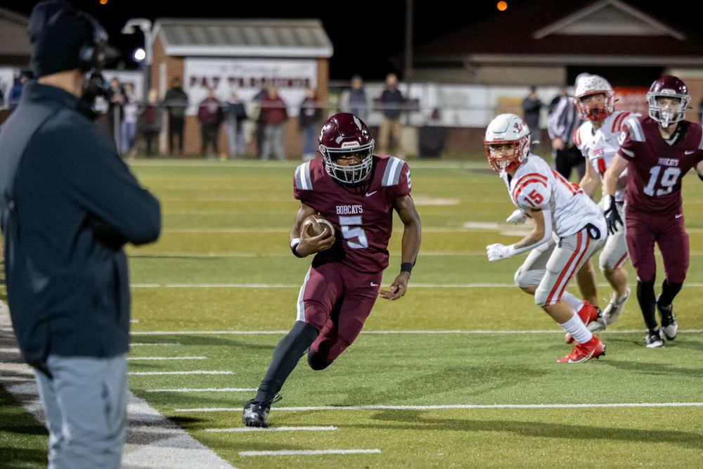 High school football player in maroon jersey runs with the ball while opponents in white uniforms pursue on the field.
