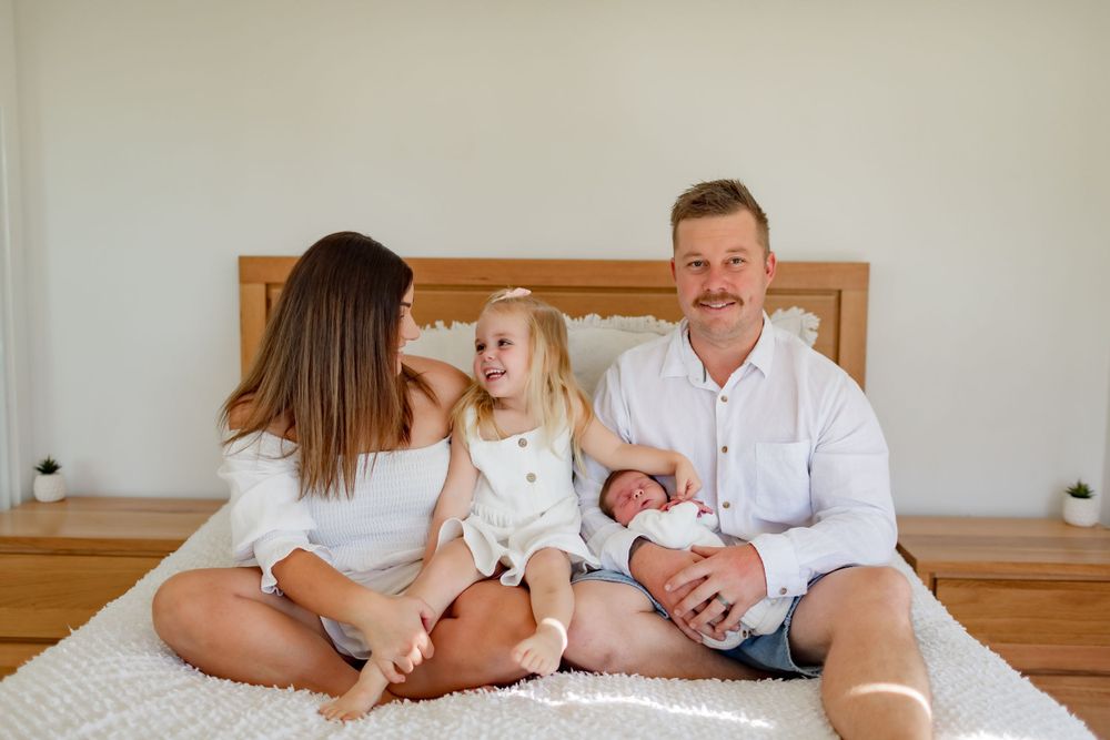 A family sits together on a bed, sharing tender moments with their newborn baby and young child.