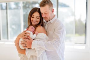 A couple holds their newborn baby wrapped in white, standing by a bright window.