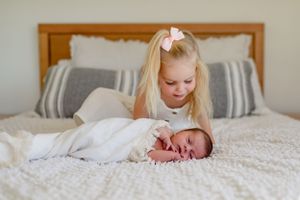 A young child looks down at a sleeping newborn baby on a white bed.