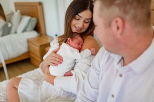 Parents gently cradle their newborn baby while sitting on a bed in a sunlit bedroom.