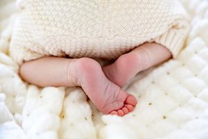 Close-up of tiny newborn baby feet peeking out from a soft cream knit blanket on a white quilted surface.