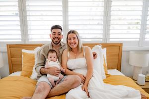 A young family relaxes together on a yellow bed with white shuttered windows behind them.