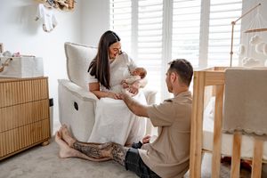 A couple sits in a nursery, with the mother holding a newborn baby in a rocking chair while the father kneels beside them.