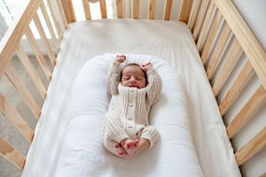 Newborn baby sleeping peacefully in a wooden crib wearing a beige knit romper on white bedding.