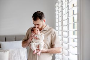 A father tenderly kisses his newborn baby while standing near bright window shutters in a serene bedroom.