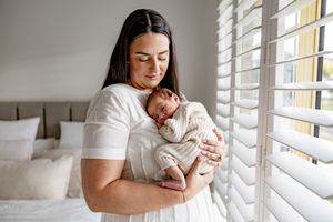 A mother in a white dress tenderly holds her sleeping newborn baby near a window with white shutters.