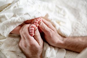 Adult hands gently cradling tiny newborn baby feet wrapped in a soft white knit blanket.