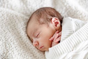 Peaceful newborn baby sleeping on a soft white knit blanket, wrapped in a white outfit with tiny hands near face.