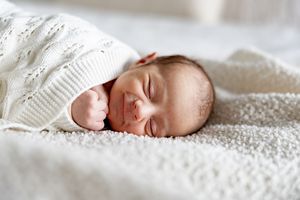 Peaceful newborn baby sleeping wrapped in a white knit blanket on a soft white surface.
