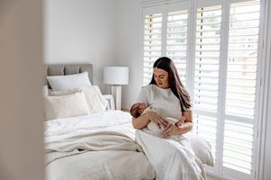 Mother in white dress breastfeeding newborn baby on bed in bright minimalist bedroom with shuttered windows.