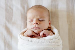 Peaceful newborn baby swaddled in white knit blanket sleeping with tiny hands tucked under chin.