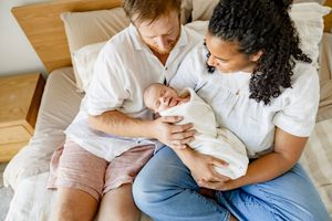 Couple sitting on bed lovingly holding their newborn baby wrapped in white swaddle blanket.
