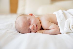 Peaceful newborn baby sleeping on white bed, wrapped in soft white blanket, eyes closed in serene slumber.