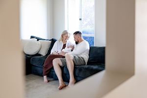Couple sitting on dark sofa at home lovingly looking at their newborn baby by window.
